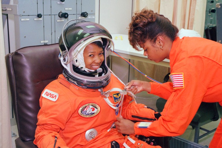 Dr Mae Jemison is a Black woman. She sits and smiles while another Black woman in a NASA uniform arranges wires on her suit